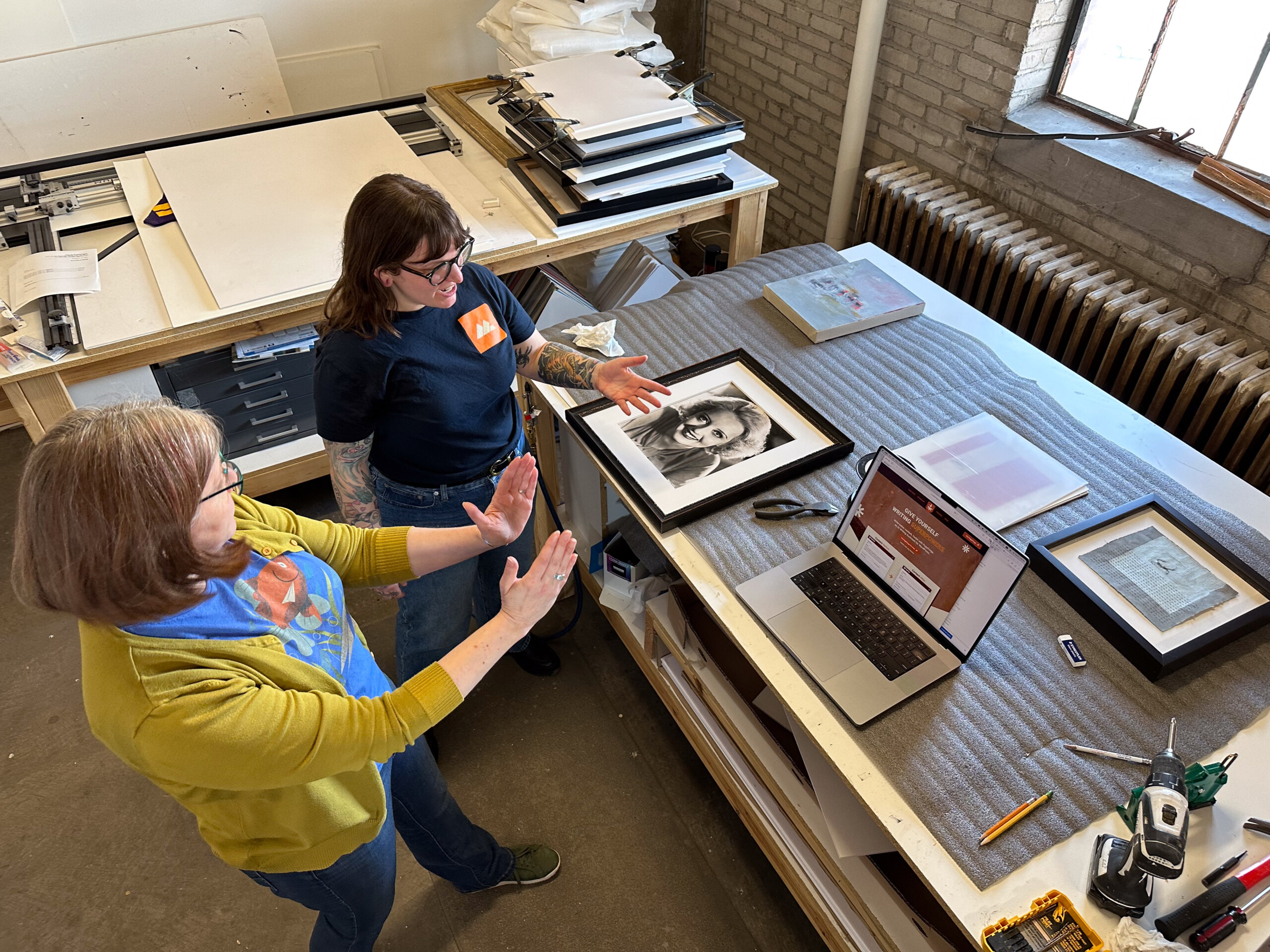 Two women in a workshop, one in a "Modern Logic" t-shirt with tattoos, and the other in a colorful shirt with a yellow cardigan, discussing over a table with a framed photo, laptop, and art supplies.