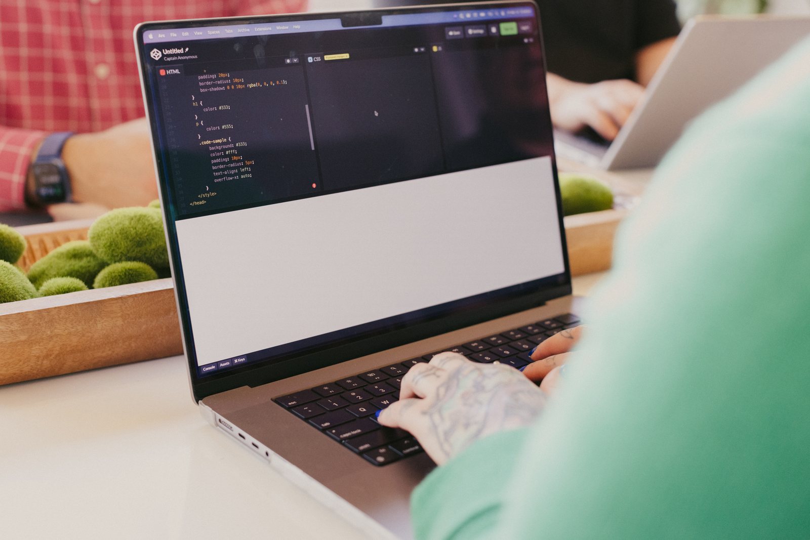 Over-the-shoulder view of a developer in a green sweater typing on a laptop with code and a browser preview open, coworkers blurred in the background